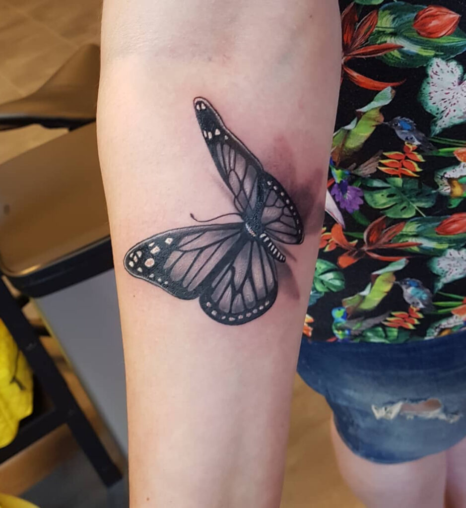 Realistische tattoo van een zwarte vlinder op de bovenarm, met lichte schaduw erachter