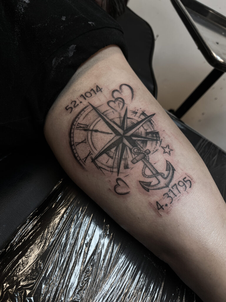 Zwart-grijze tattoo met kompas, anker, harten en coördinaten op de arm, gezet bij Tat2holics in Den Haag