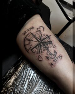 Zwart-grijze tattoo met kompas, anker, harten en coördinaten op de arm, gezet bij Tat2holics in Den Haag