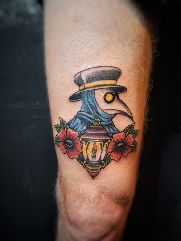 Tattoo van een plaagdokter met hoed en snavelmasker boven een lantaarn en bloemen, in kleurrijke neo-traditionele stijl op het onderbeen, geplaatst bij Tat2holics in Den Haag