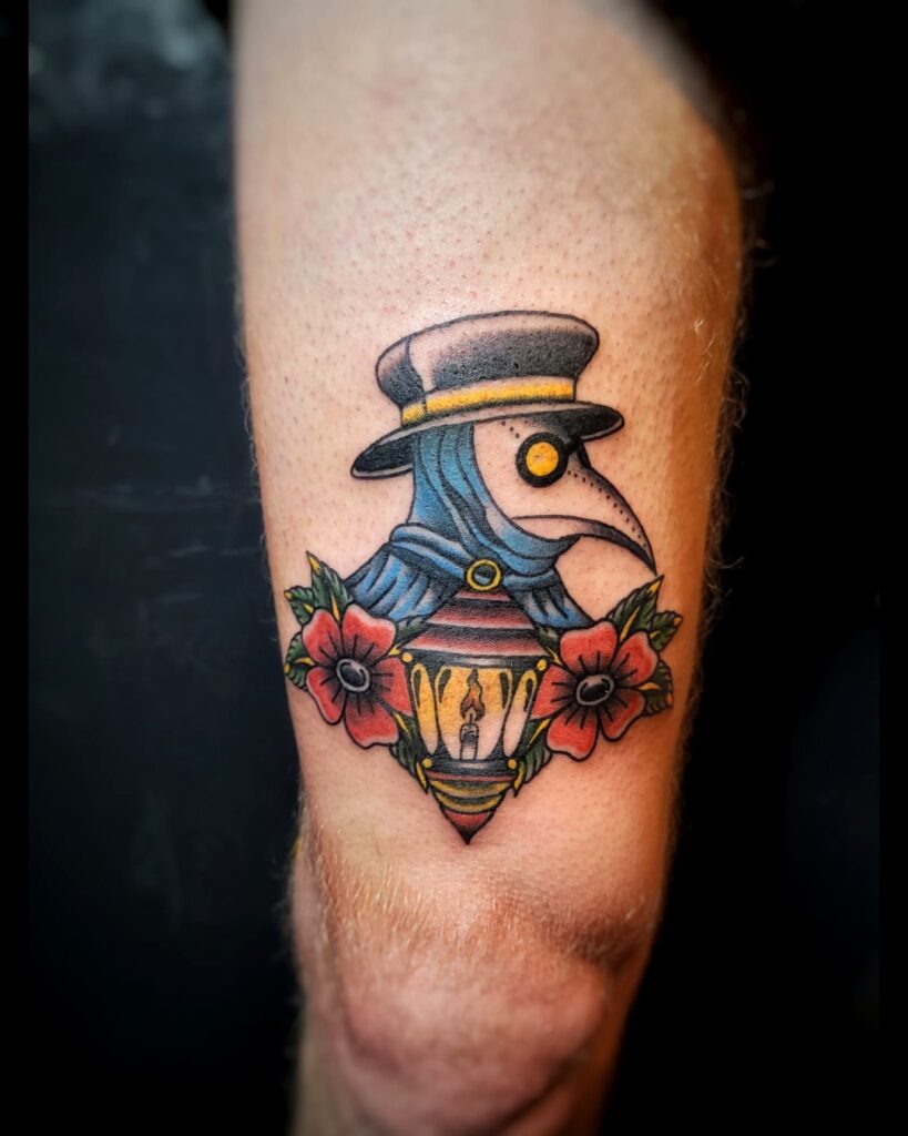 Tattoo van een plaagdokter met hoed en snavelmasker boven een lantaarn en bloemen, in kleurrijke neo-traditionele stijl op het onderbeen, geplaatst bij Tat2holics in Den Haag