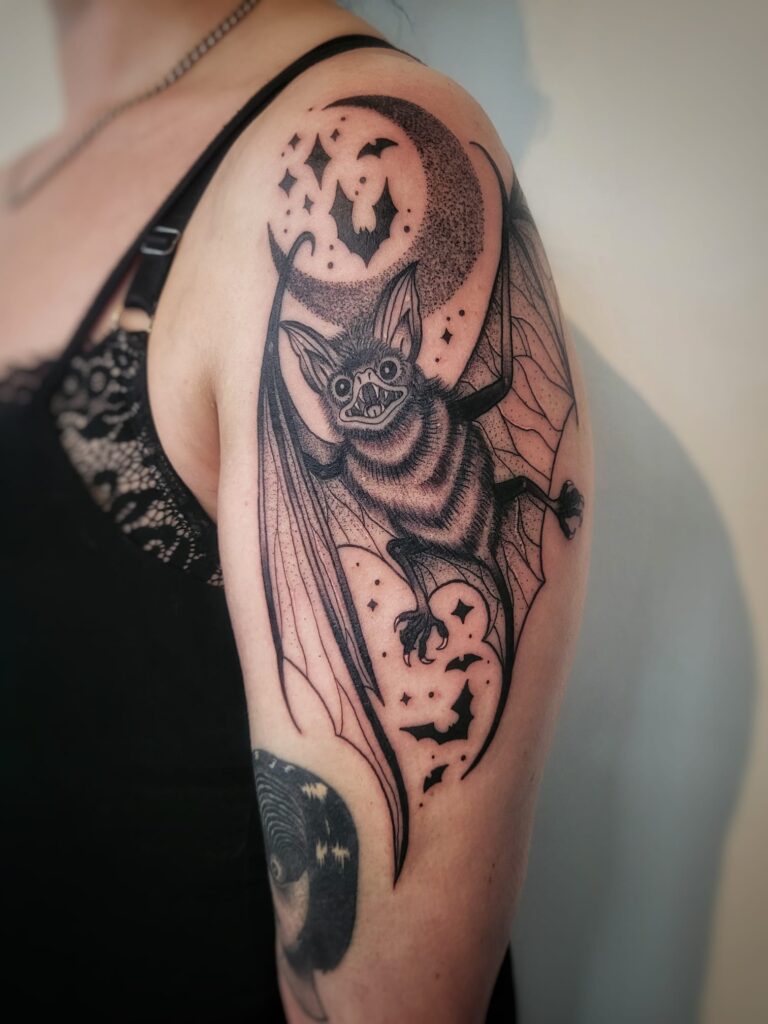 Tattoo van een realistisch getekende vleermuis met gespreide vleugels, zwevend voor een halve maan met sterren en vliegende silhouetten, op de bovenarm van een vrouw