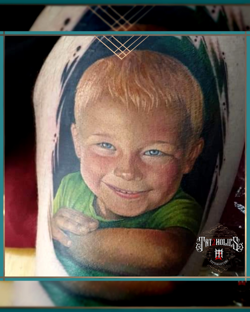 Tattoo van een blond jongetje met blauwe ogen en groene shirt, levensecht uitgewerkt in kleur op de arm, geplaatst bij Tat2holics in Den Haag