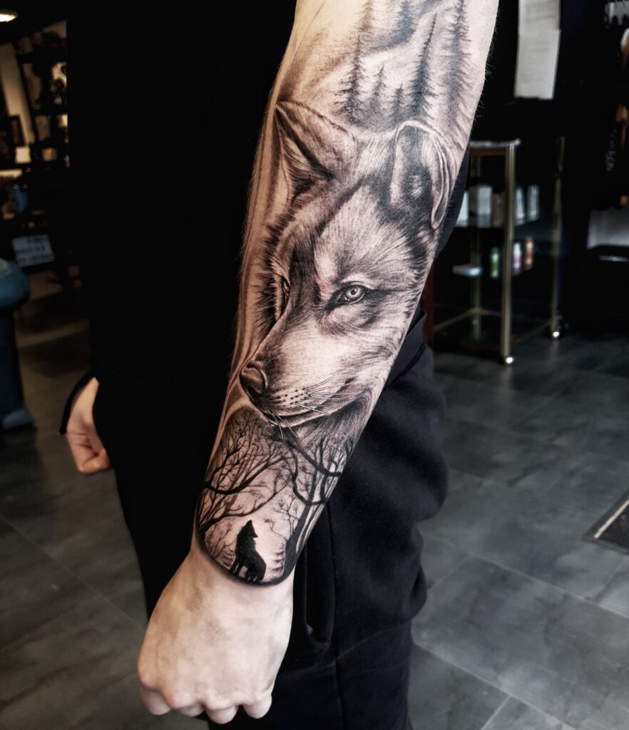 Realistische tattoo van een wolf met intens oogcontact, geplaatst op de onderarm, met een bos en huilende wolf in silhouet