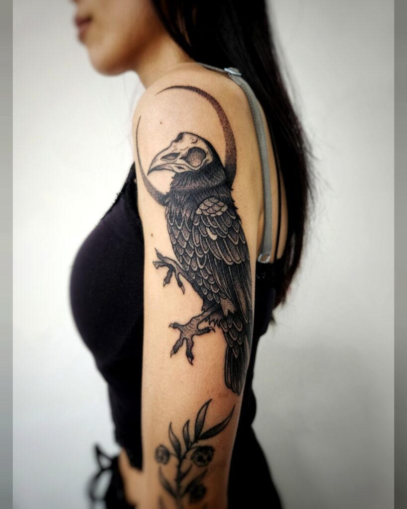 Tattoo van een zwarte raaf met gedetailleerde veren, zittend onder een donkere maansikkel, geplaatst op de bovenarm van een vrouw