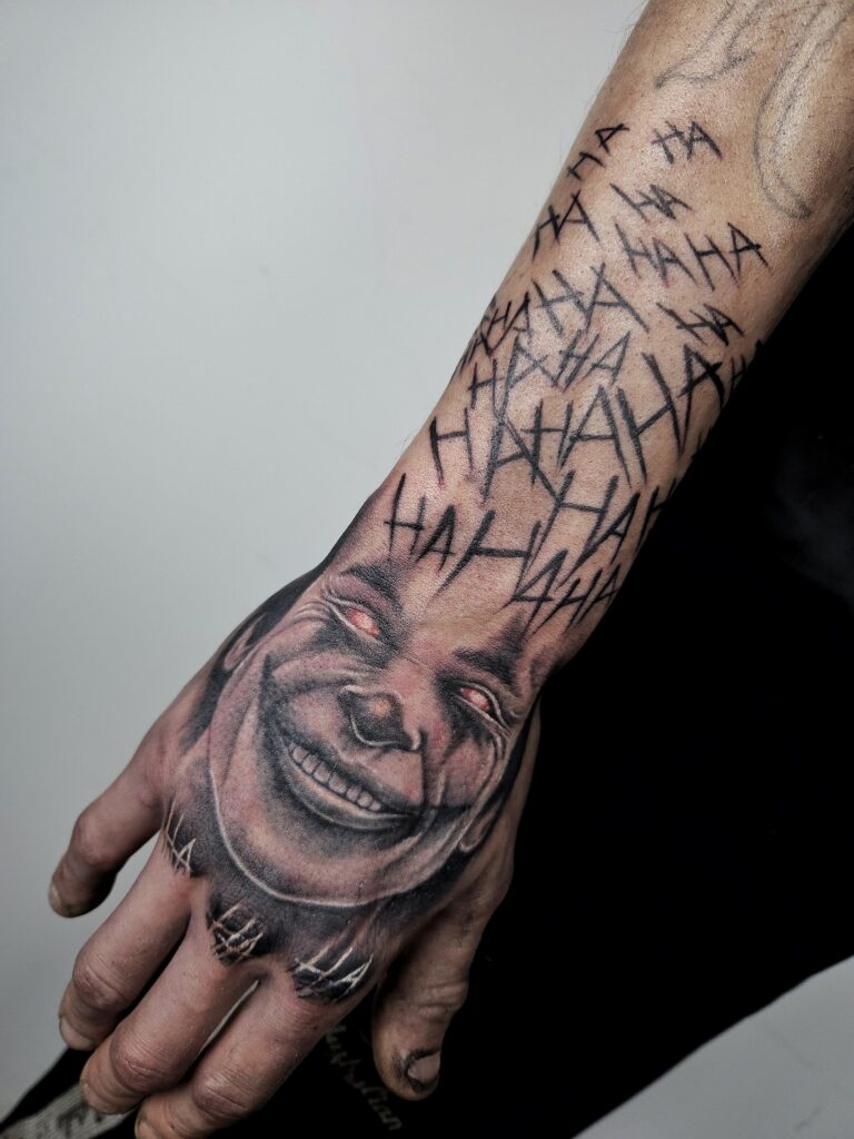 Tattoo van een angstaanjagende Joker op de hand, omlijst met ‘HAHA’ teksten in verschillende groottes