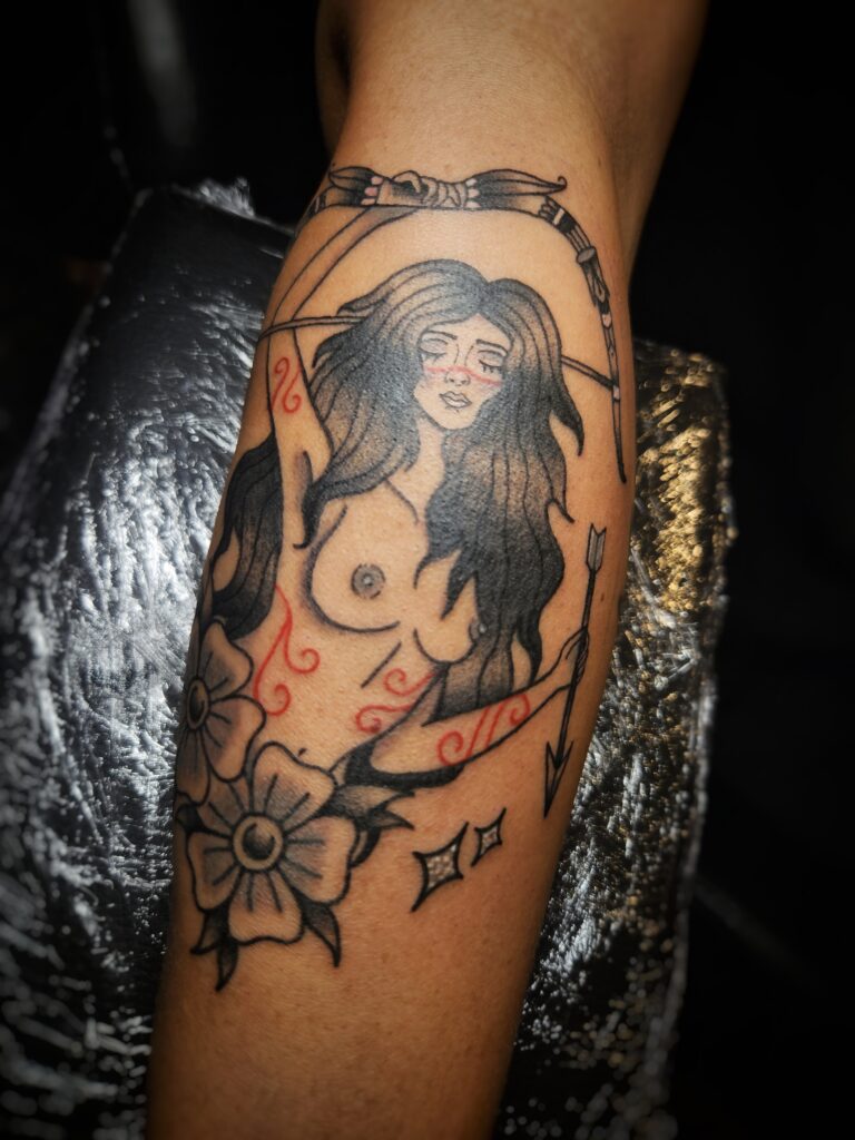 Tattoo van een naakte vrouwelijke boogschutter met zwarte haren, bloemen en pijl en boog in neo-traditionele stijl, gezet op de arm bij Tat2holics in Den Haag