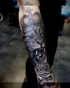 Tattoo van een brullende leeuw met kroon en roos op de onderarm, zwart-grijs realisme